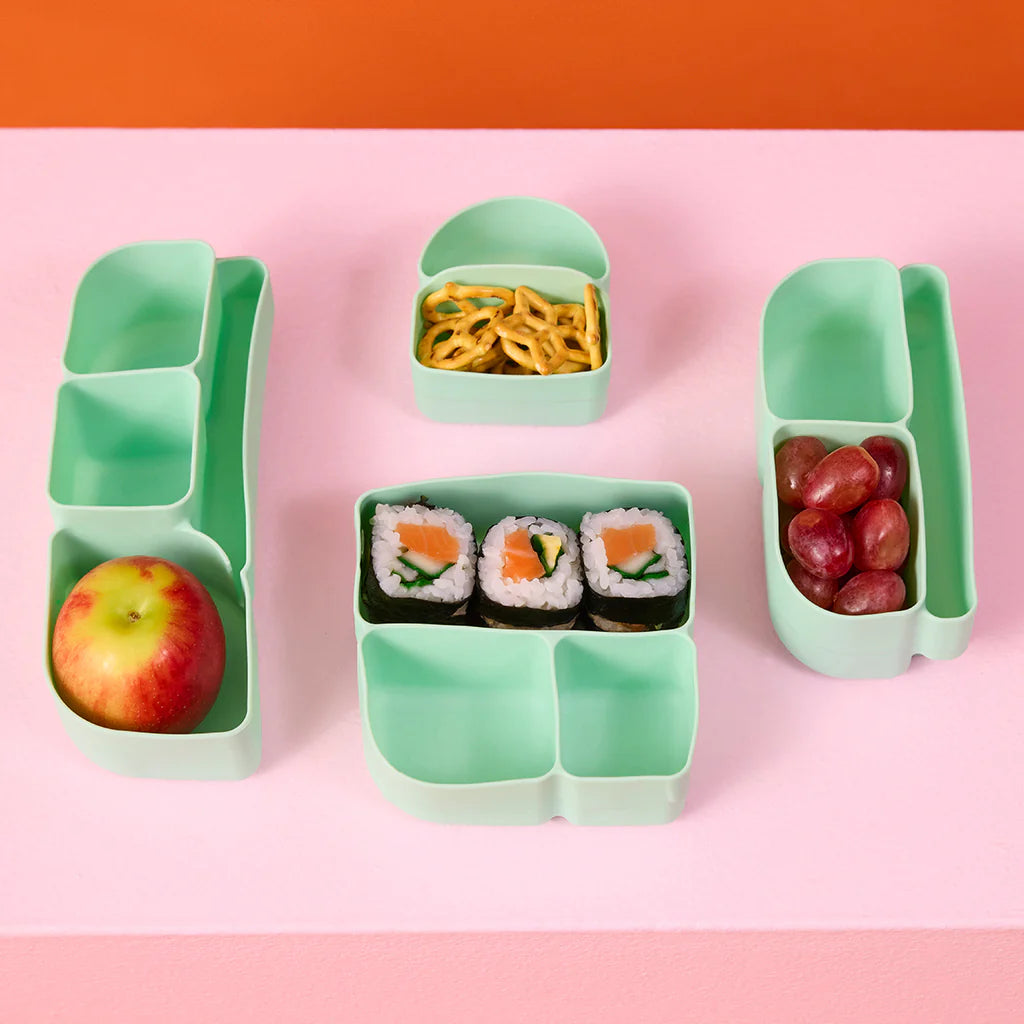 Forest green bento box with compartments containing an apple, sushi, and grapes on a pink background.