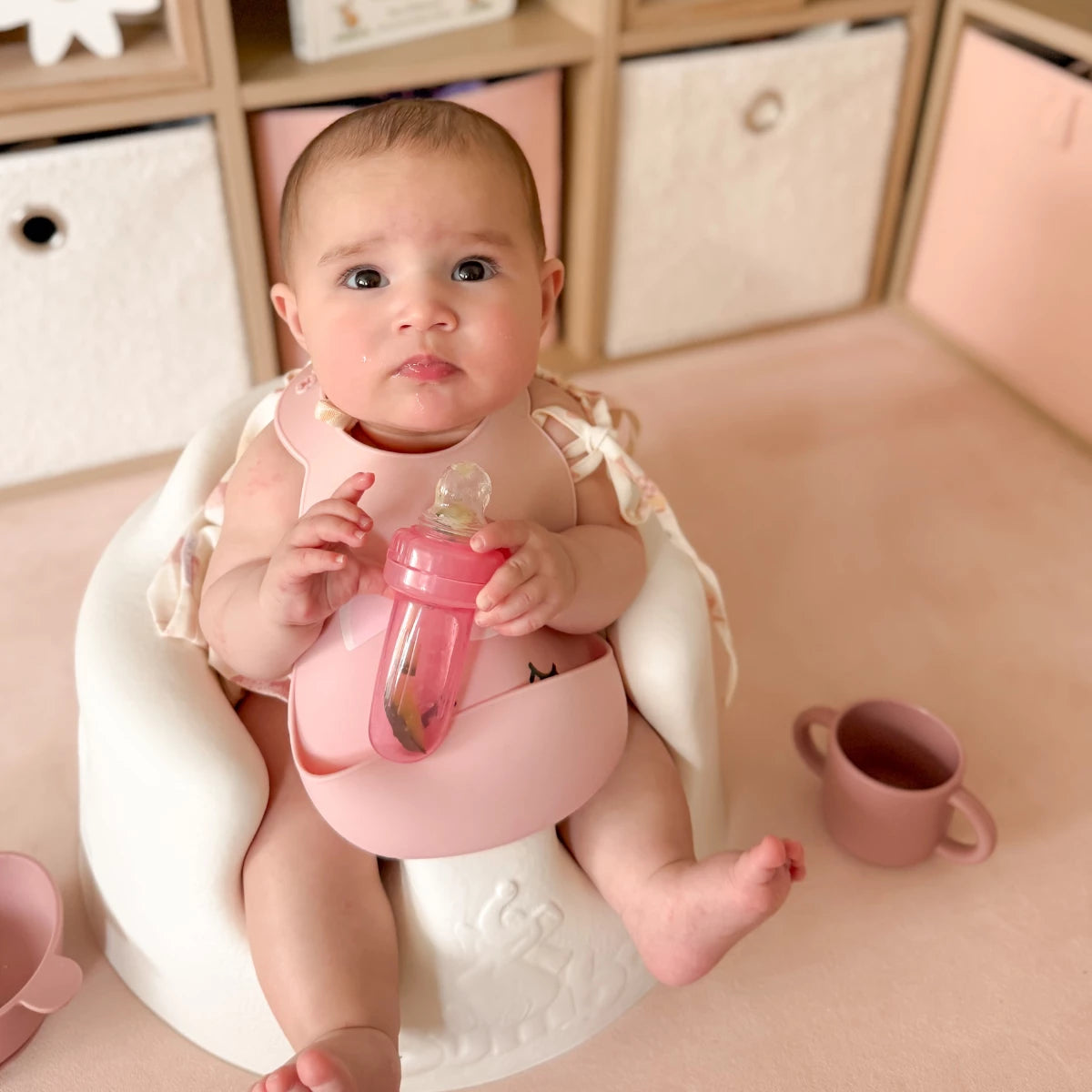 A baby sitting in a cotton bumbo floor seat, wearing a pink bib and holding a pink bottle, with a matching pink cup on the floor nearby.