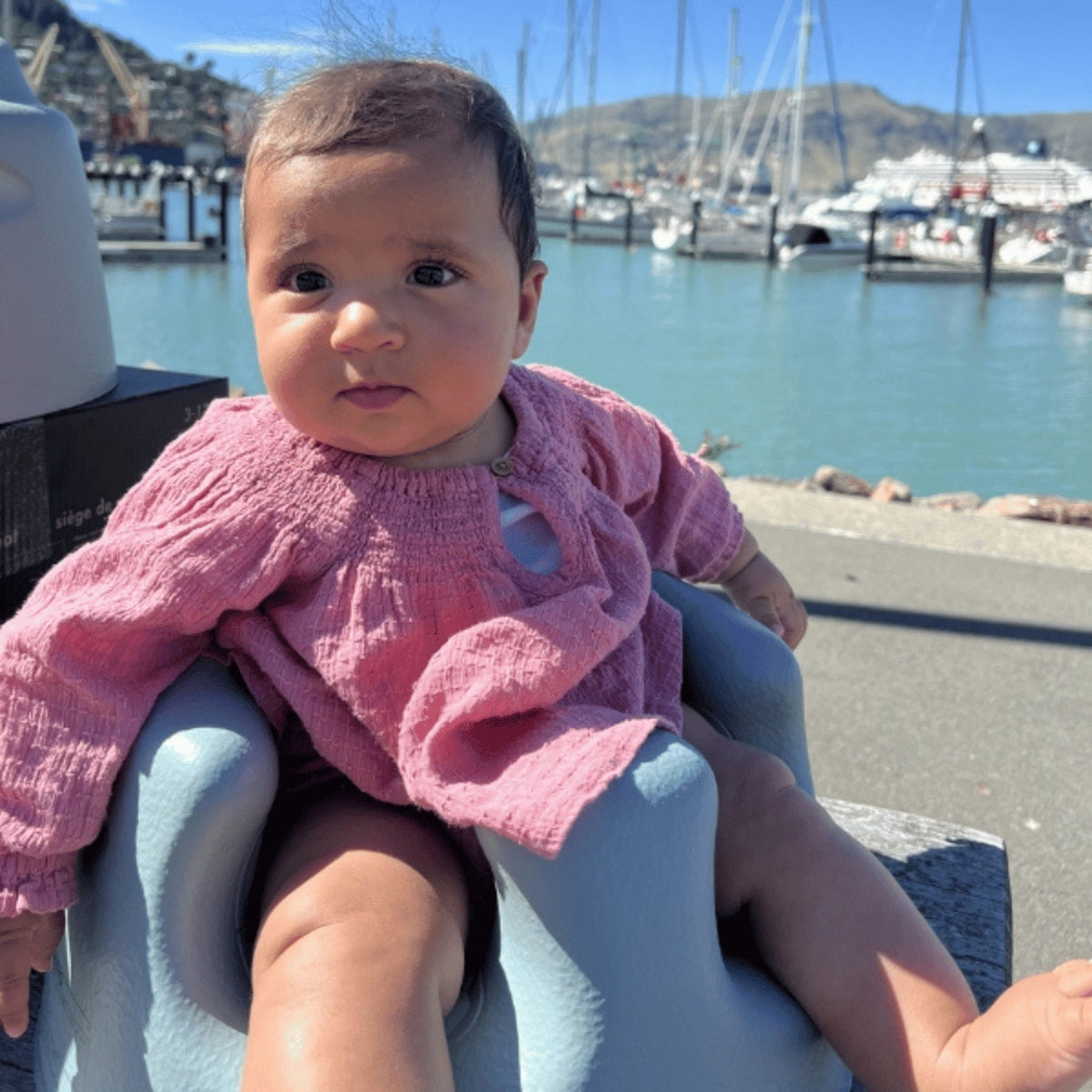Baby sitting upright in a Saltbush Bumbo Floor Seat Max during an outdoor day trip by a marina harbor