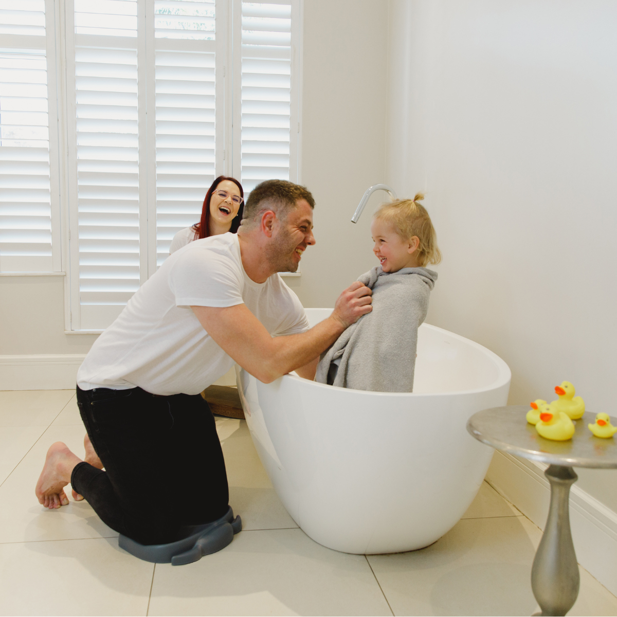 A father kneels beside a white freestanding bathtub using Bumbo kneeling pad slate gray and a young child sits inside wrapped in a gray towel. A woman stands in the background laughing near white shutters, and small yellow rubber ducks sit on a side table.