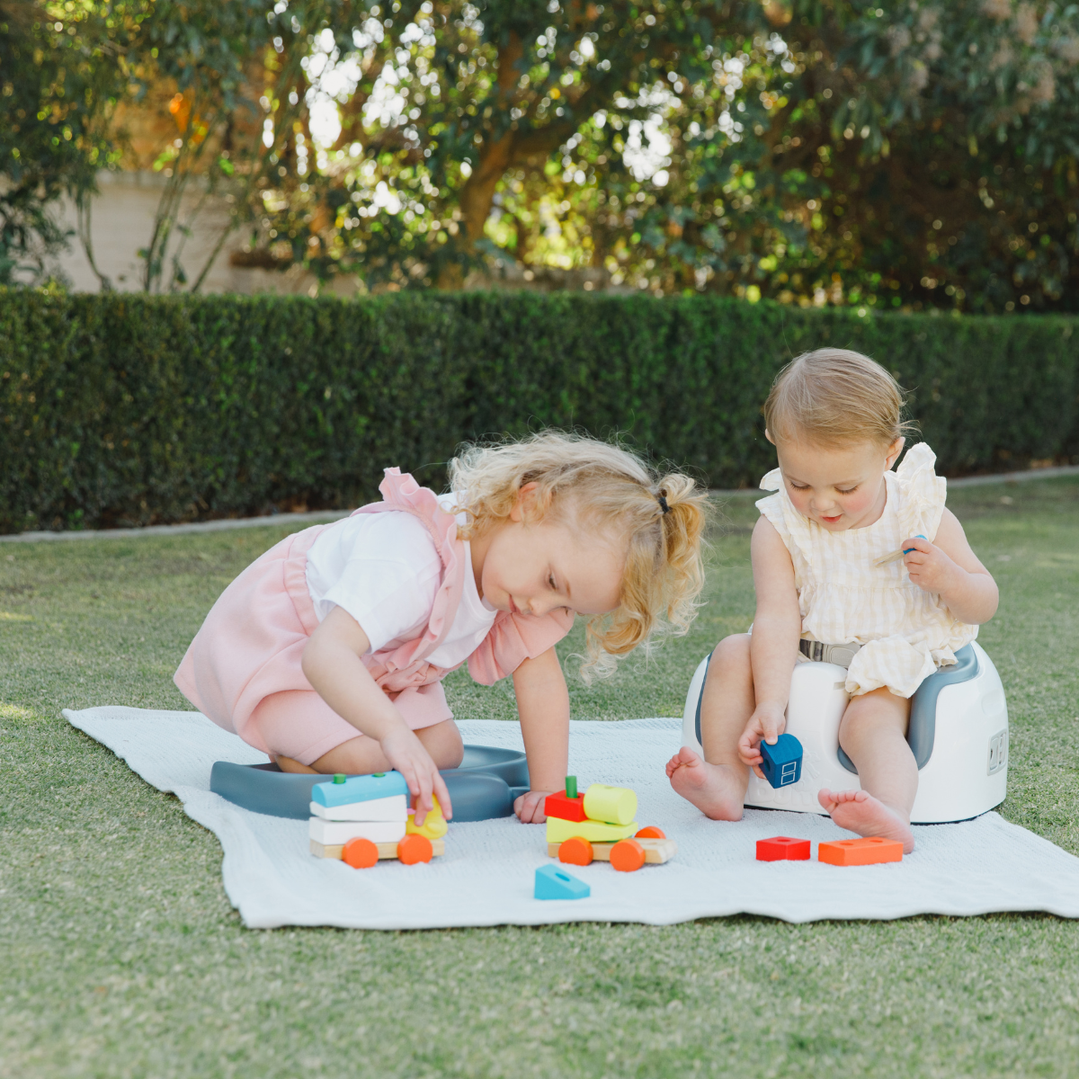 Two young children playing outdoors on a white blanket on a grassy lawn. The child on the left wears a pink and white outfit and is kneeling Bumbo kneeling pad while playing with colorful toy blocks, and the child on the right sits on a bumbo 3in1 multi-seats  wearing a cream-colored outfit.