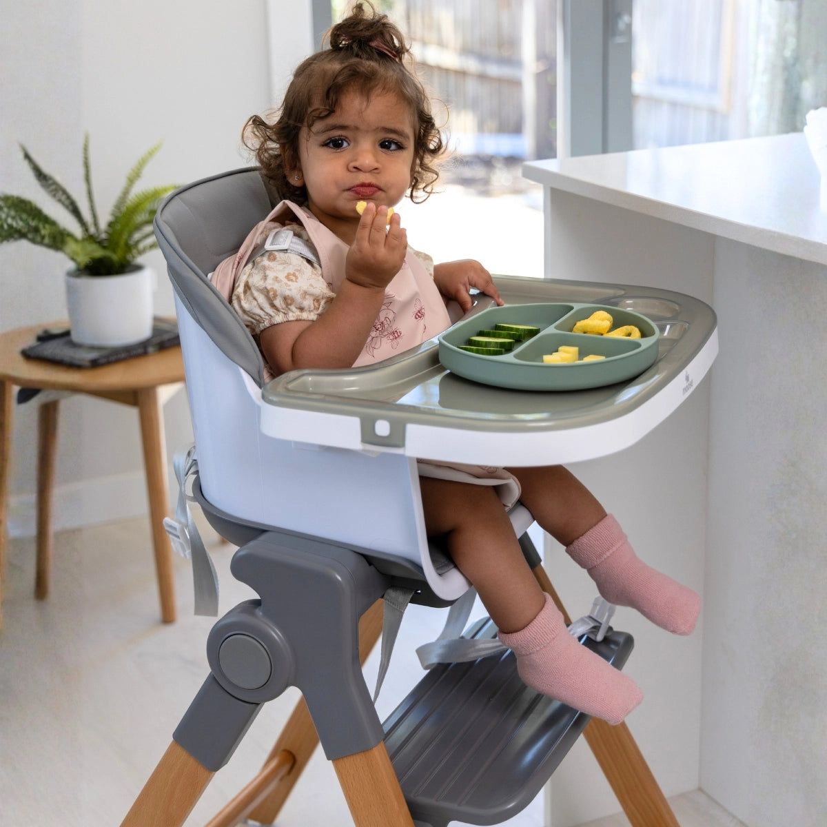 A toddler sits in a Frankie Foldable high chair with a tray in a bright indoor room. The high chair is light gray and white with wooden legs, and the tray holds green beans and yellow food pieces.