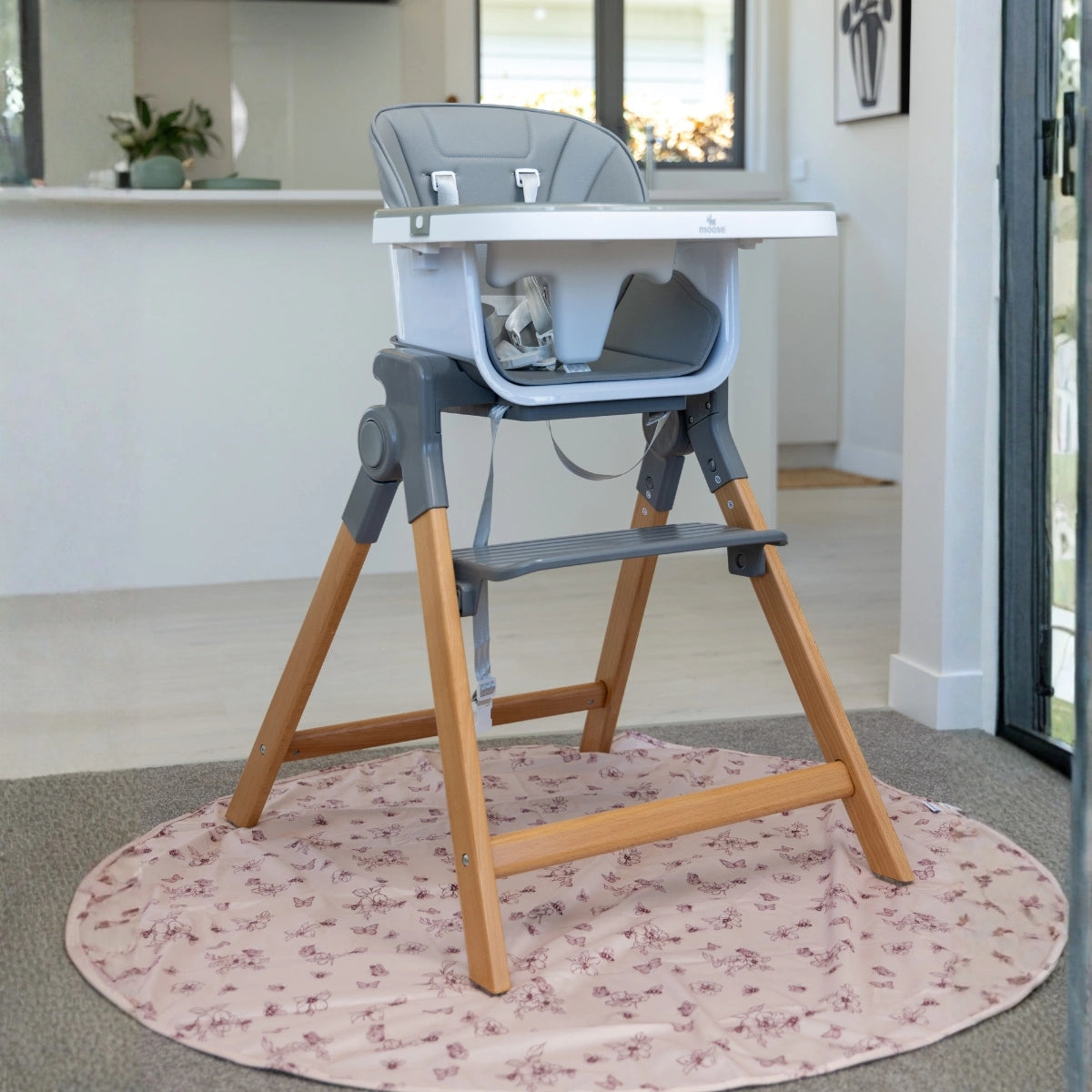 Frankie high chair with a white tray and light gray seat, supported by natural wood legs, placed on a round pink floral rug in a bright indoor room