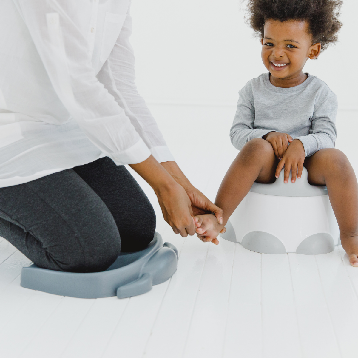A child sitting on a white potty with gray accents while an adult kneels in a Bumbo kneeling pad slate gray beside them, holding the child's hand. The adult is wearing a white long-sleeve top and dark gray leggings, and the child is wearing a light gray long-sleeve shirt.