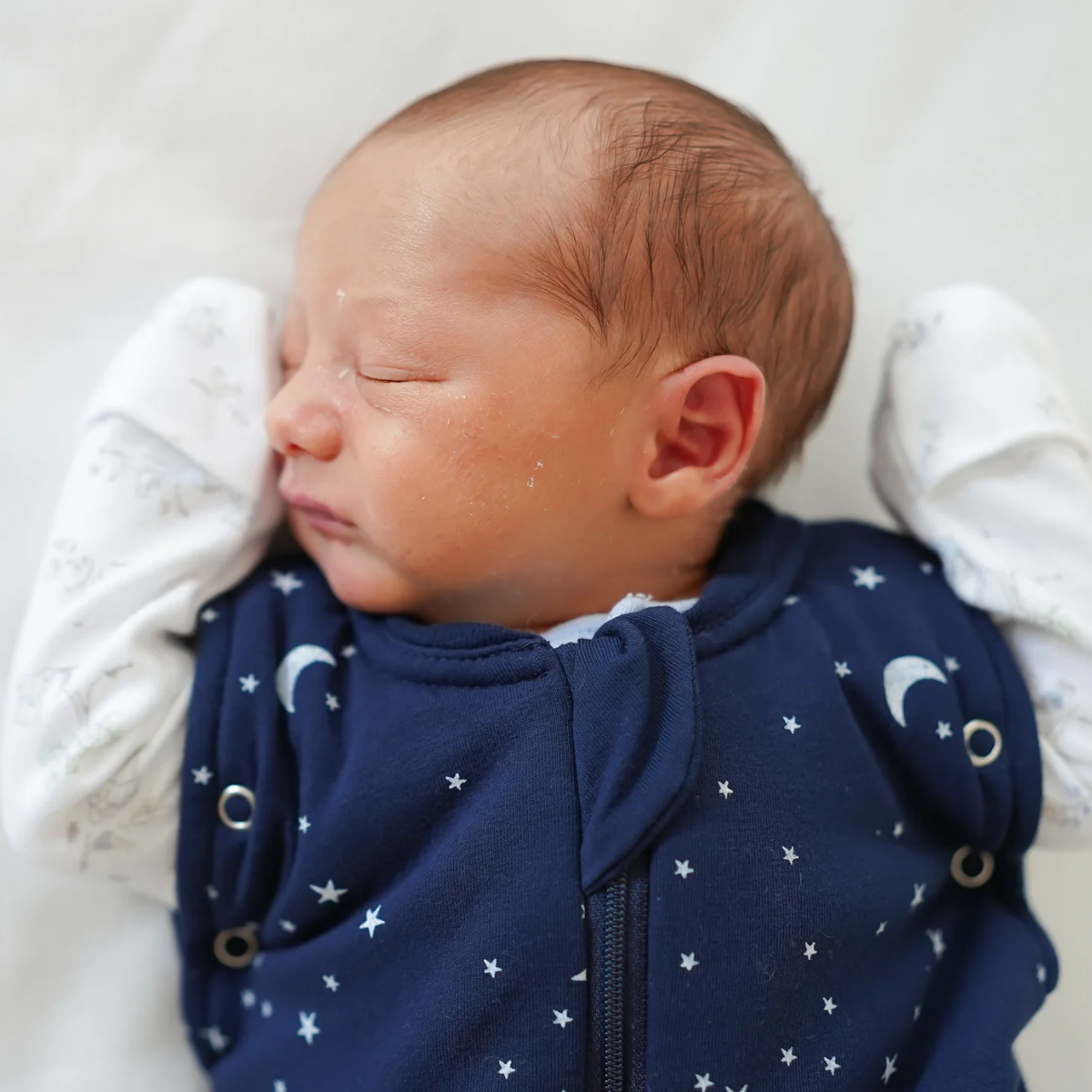 A sleeping newborn baby wearing a Sweet Dreamers Sleep & Swaddle Transition Bag with a white star and moon pattern, tucked in white blankets.