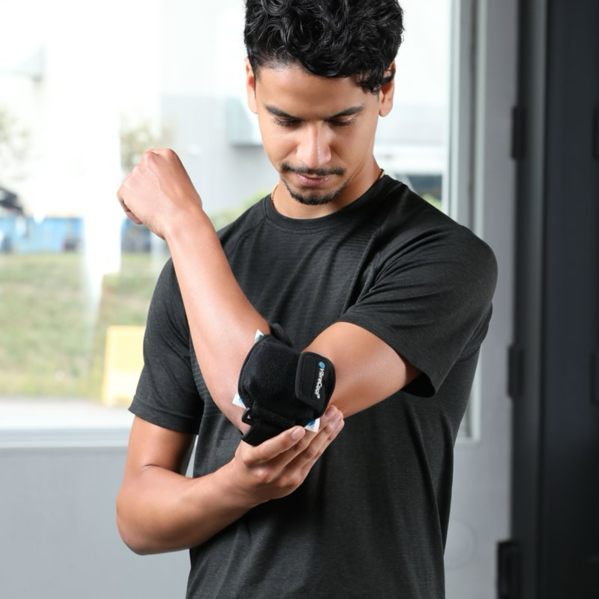 A man wearing a black athletic t-shirt demonstrates the VibraCool Easyfit, elbow support brace on his forearm in an indoor setting.