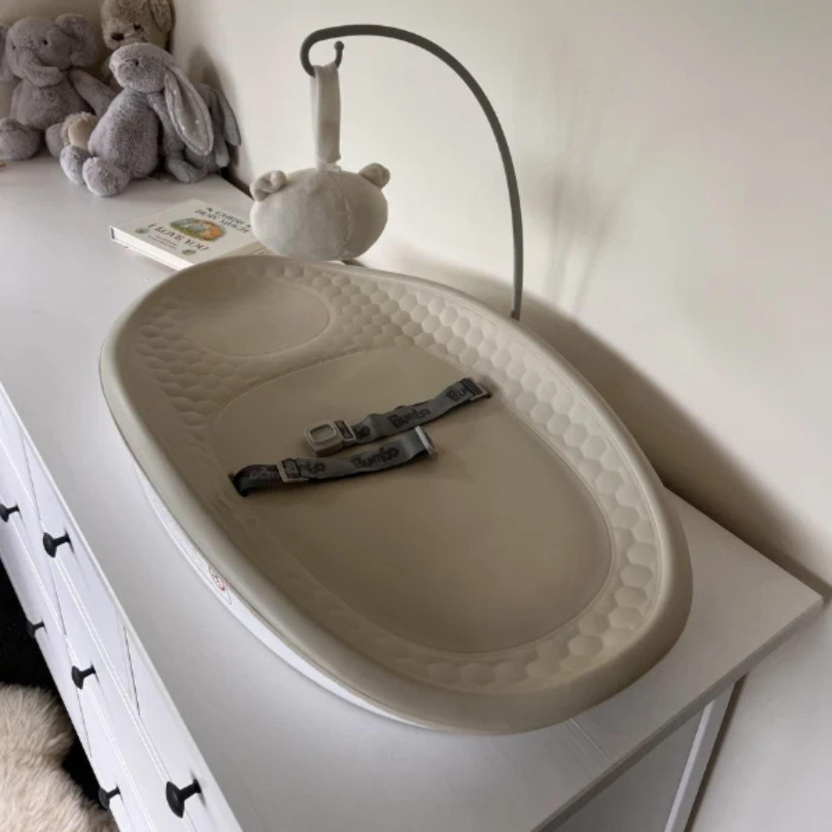 Bumbo change pad max in a taupe color, resting on a white dresser. It is shown with its matching fabric safety strap and a small plush toy attached to a hanging stand.