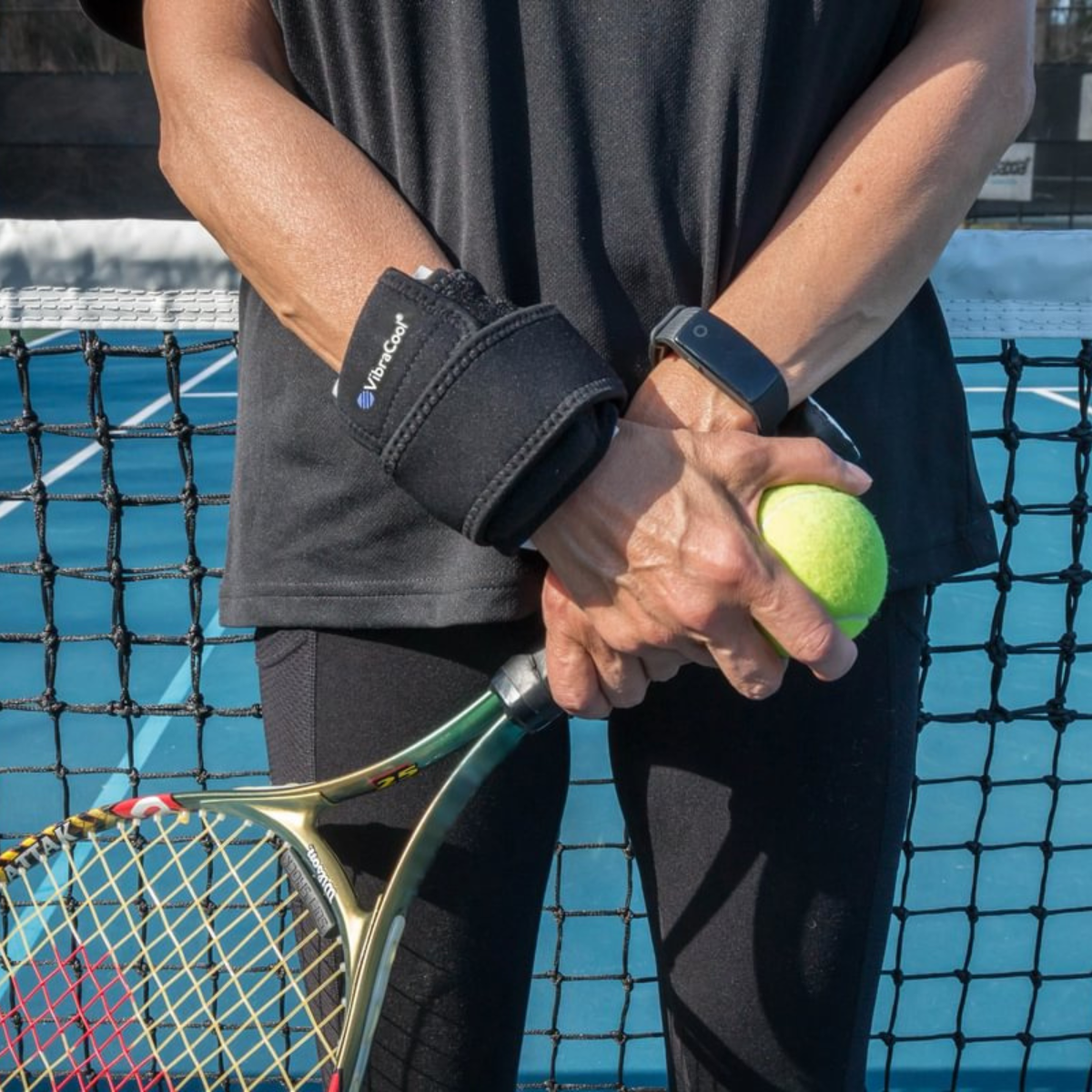 A close-up of a person on a tennis court wearing a VibraCool Easyfit on one wrist and holding a tennis racket and tennis ball. The brace has a white logo that reads 'TENSER'.