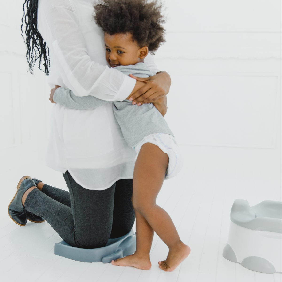 A woman kneels on a Bumbo kneeling pad while holding a young child who is wearing a light gray long-sleeve top and diaper. A matching gray potty seat is visible on the floor to the right in a bright, white indoor setting.