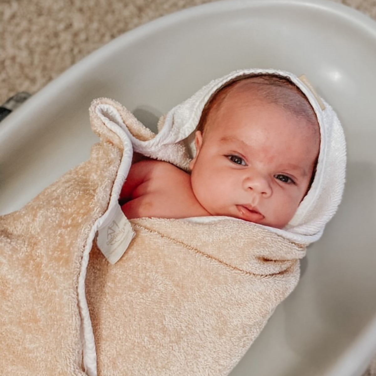 A newborn baby wrapped in a cuddle dry towel and a white hooded towel, lying inside a white bathtub.