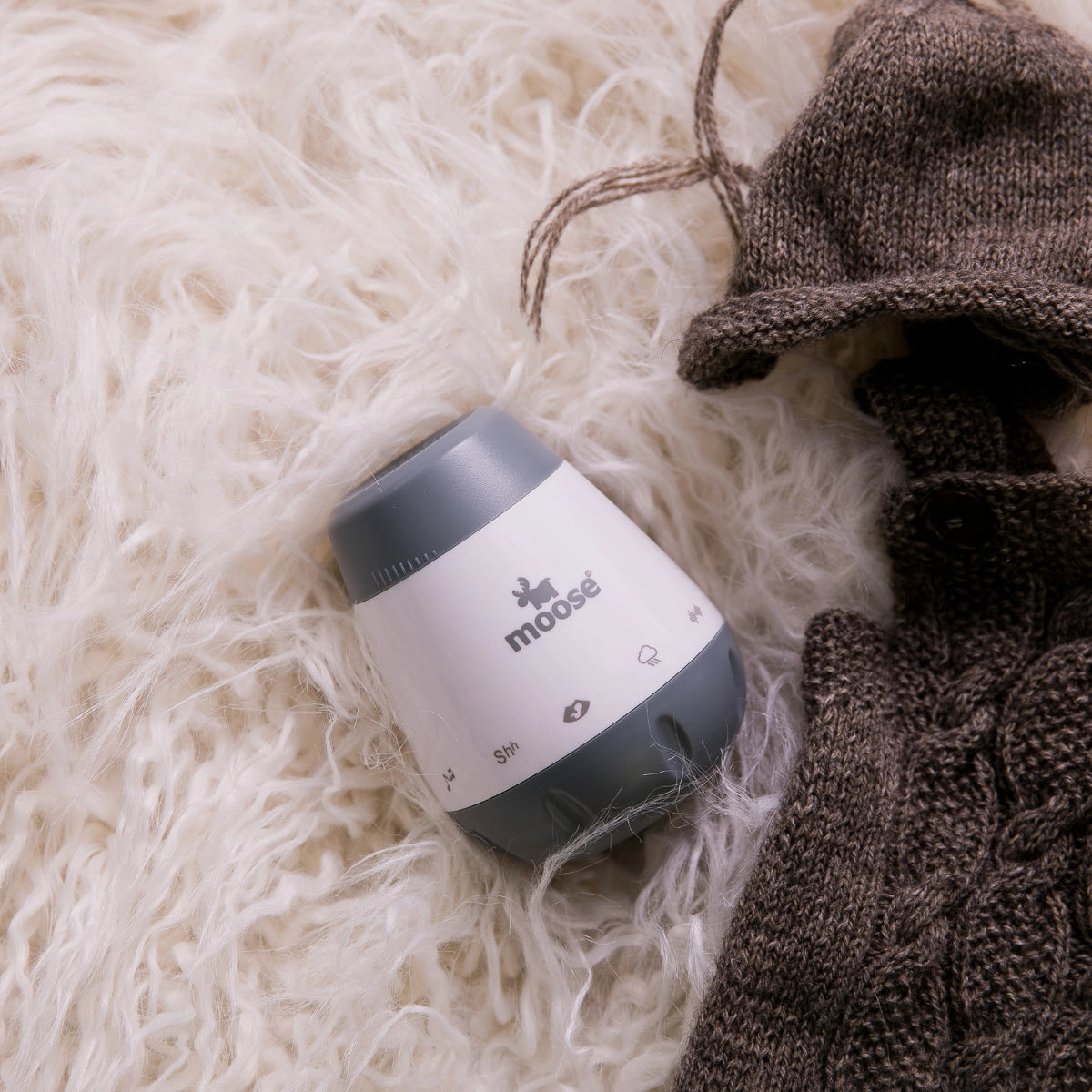 Grey and white Calm-a baby white noise machine on a soft blanket beside knitted baby clothes