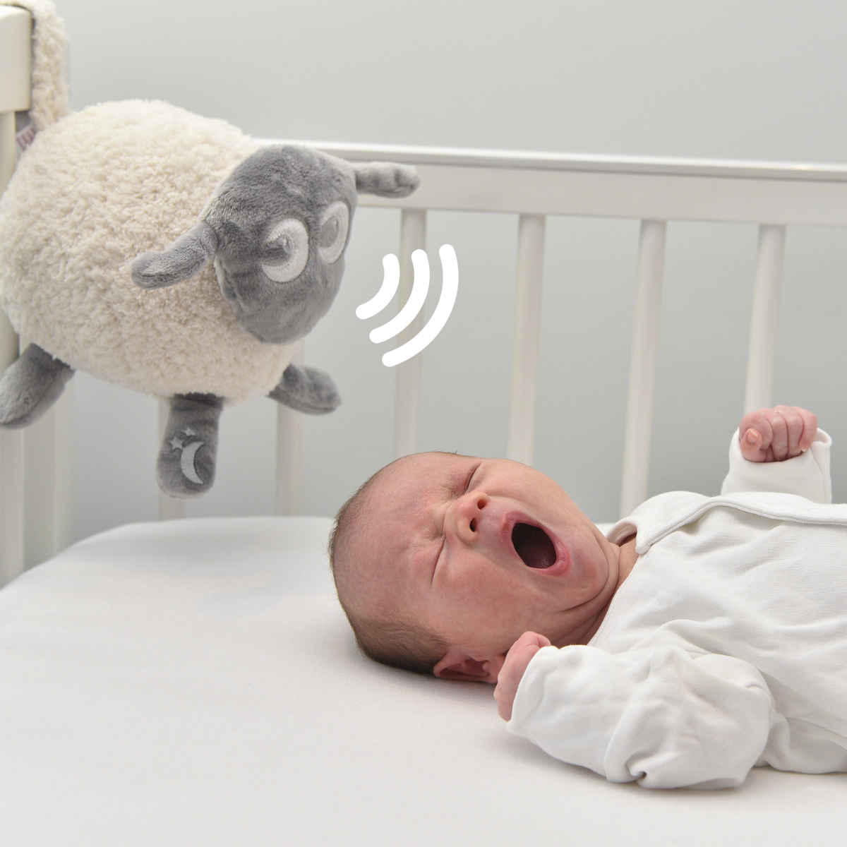 A newborn baby wearing a white onesie lies in a white crib while a DELUXE Ewan the Dream Sheep with a sound wave graphic floats above, suggesting a baby monitor or sound alarm product.