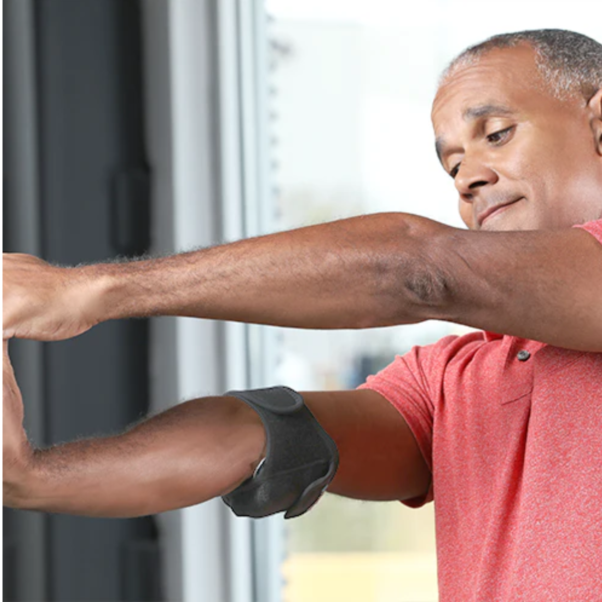 A man wearing a red polo shirt demonstrates the use of a VibraCool Easyfit on his forearm, with a window in the background.