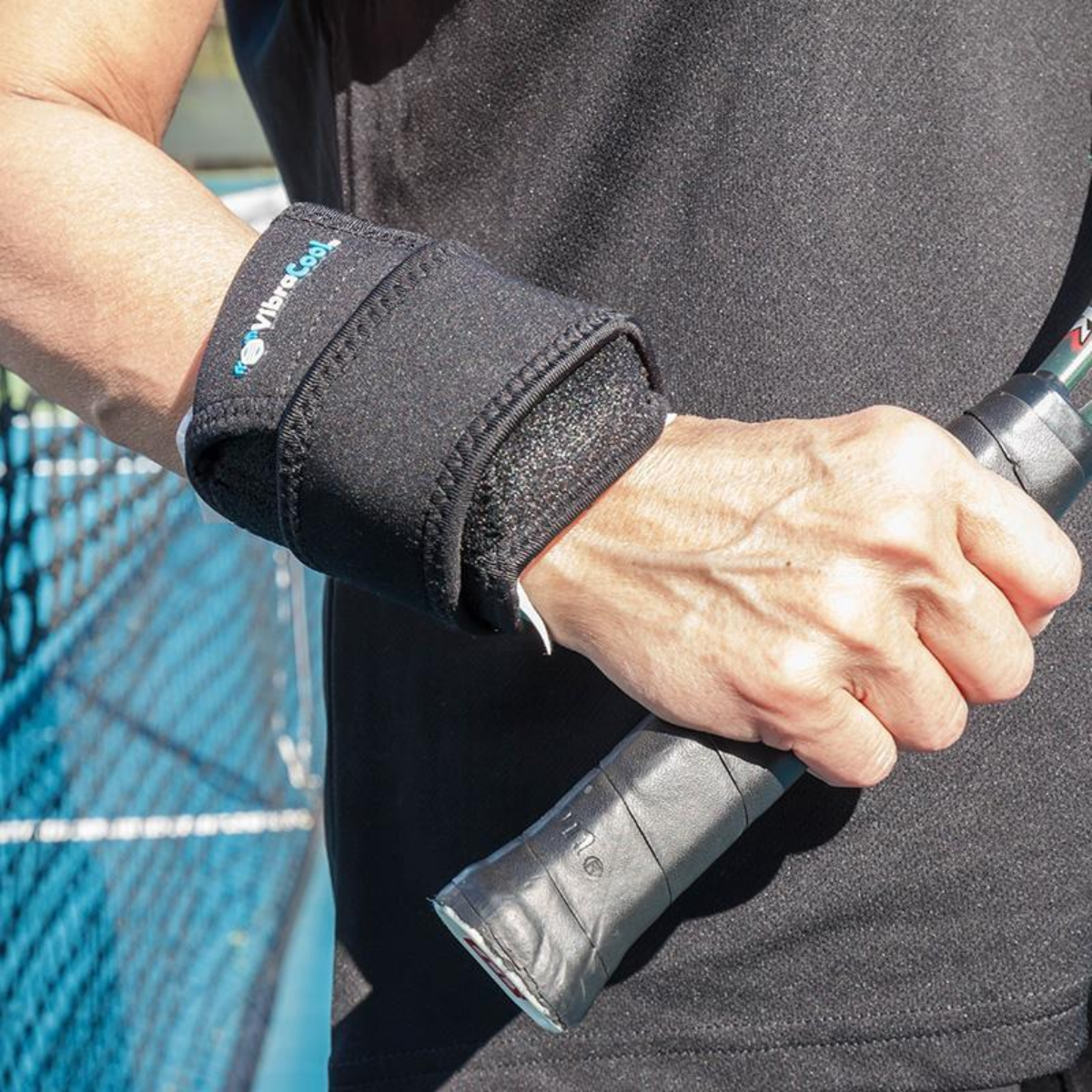 A close-up of a person's hand gripping a tennis racket handle while wearing a VibraCool easyfit, support sleeve with a visible Vibram logo, set against a tennis court background.