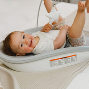A baby lying on their back in a Bumbo change pad max, smiling and reaching for a white star toy hanging above. The baby is wearing a white t-shirt and patterned shorts, and the tub sits on a textured cream blanket.