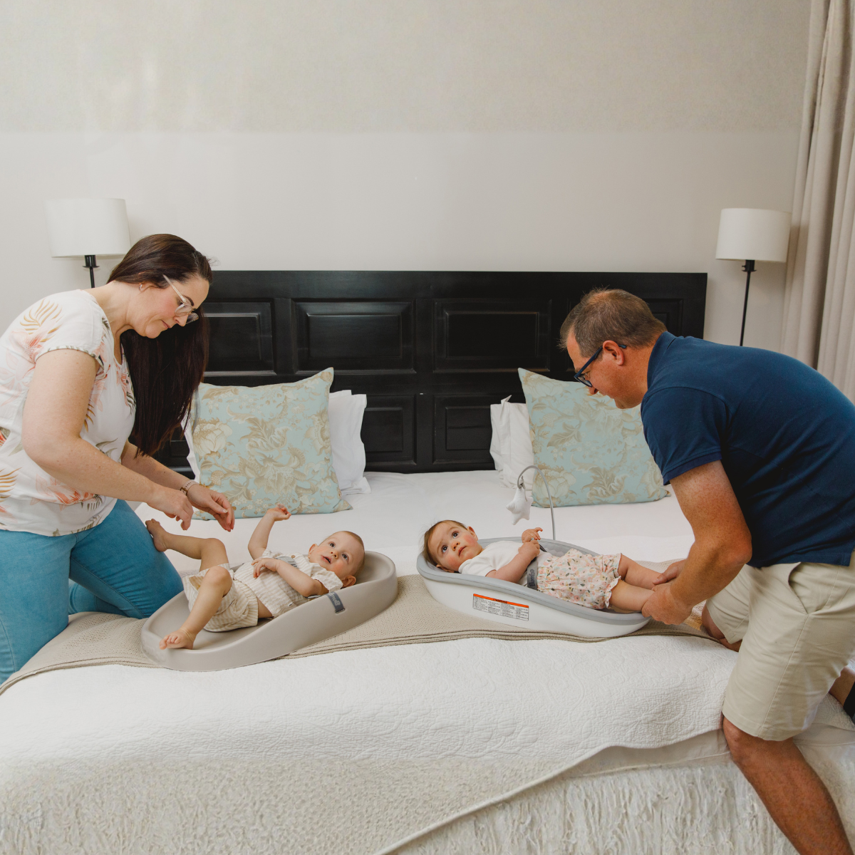 A family of four is shown in a bedroom setting, using baby changing tables on a bed. The scene features a woman on the left in a white floral top and blue jeans, and a man on the right in a navy blue polo shirt and khaki shorts, both interacting with babies lying on bumbo change pad and change pad max