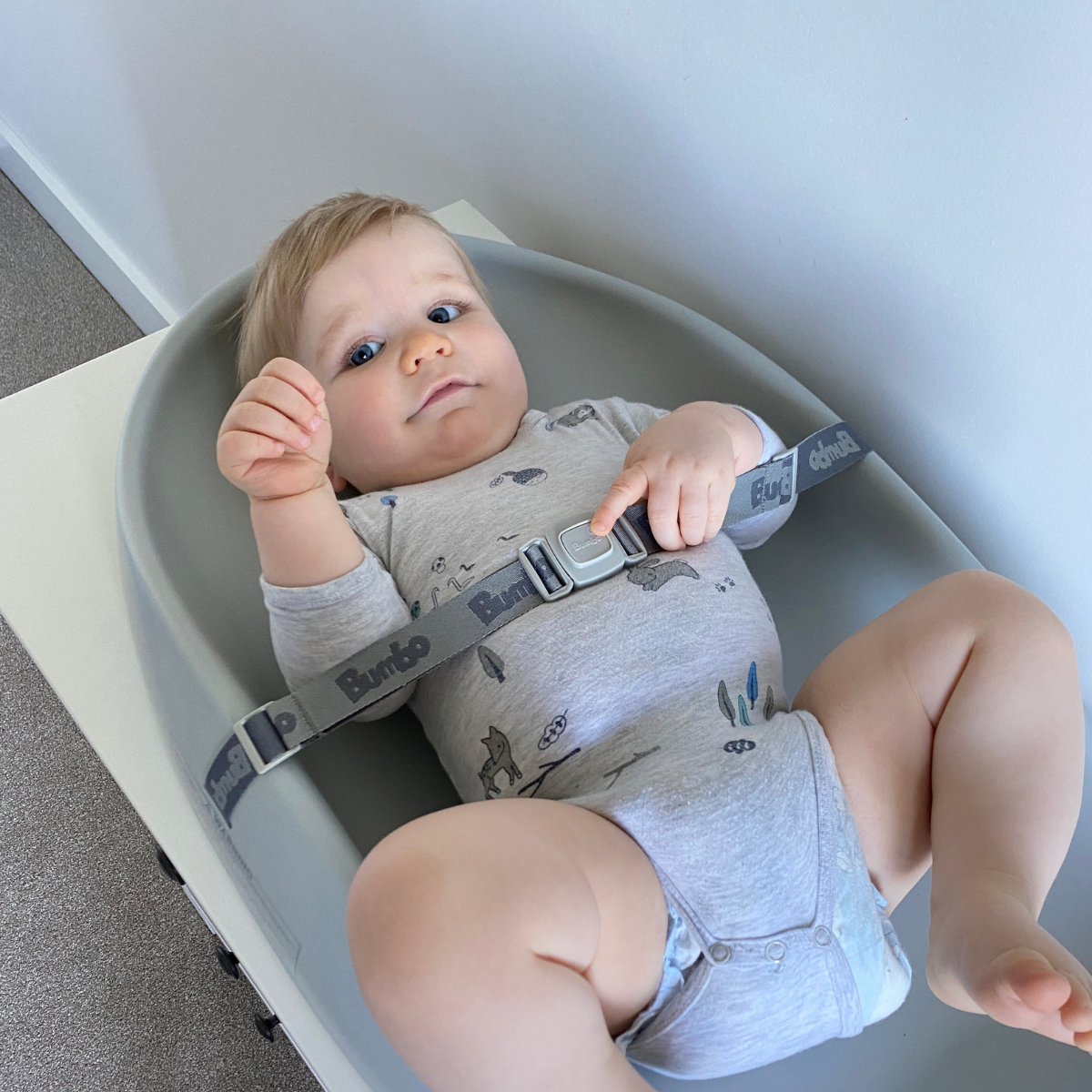 A baby lying on a gray changing pad, wearing a light gray long-sleeve romper and diaper cover with a shark print, secured by a gray shoulder harness with a Buck brand buckle.
