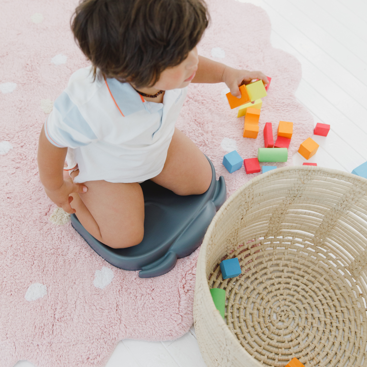A toddler kneels on a Bumbo kneeling pad  on a pink cloud-shaped rug, playing with colorful wooden blocks next to a woven basket.