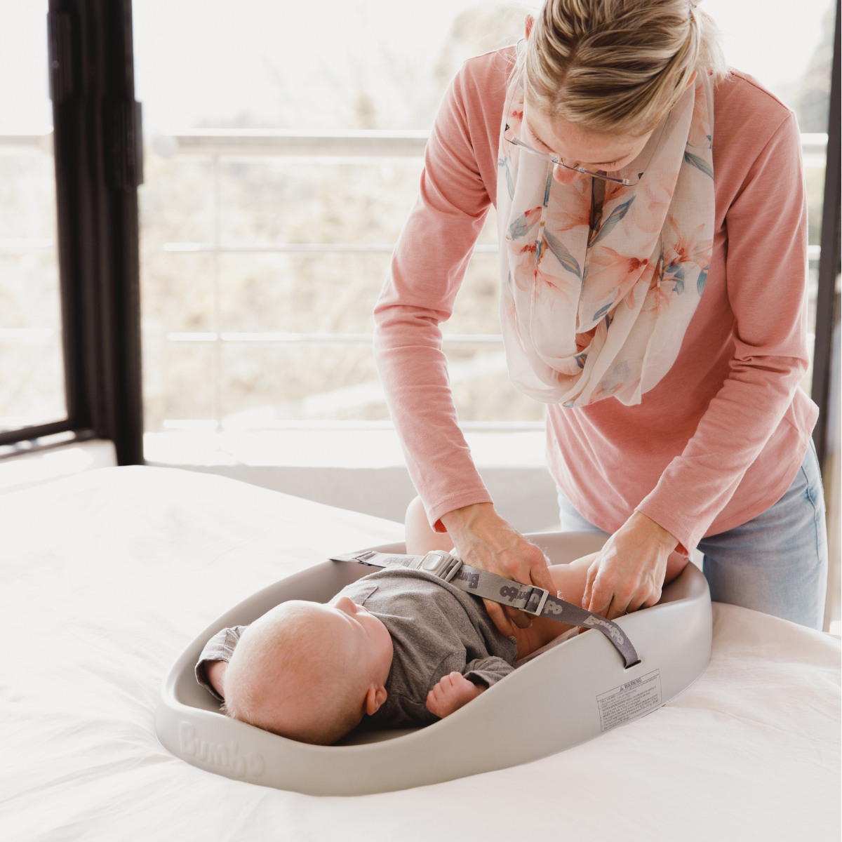 A woman in a pink long-sleeve top and floral scarf is adjusting the harness on a baby lying in a taupe changing pad baby support seat on a white changing table. The brand name 'Pajama Baby' is visible on the side of the seat.