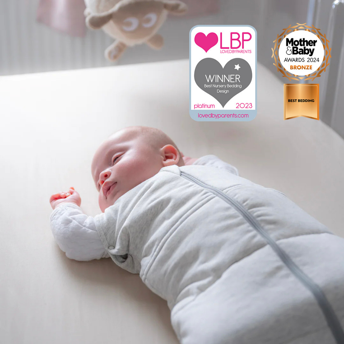 A sleeping baby wearing a light gray long-sleeve swaddle blanket lies on a cream-colored changing table. Two award badges are displayed at the top right: the LBP Platinum 2023 Winner badge and the Mother & Baby Awards 2024 Bronze Best Bedding badge.