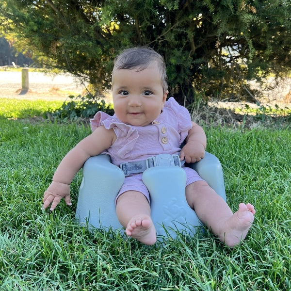 Bumbo Floor Seat Max in Saltbush with wider leg openings and 3-point harness supporting a smiling baby seated on a grass