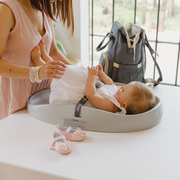 A baby changing pad scene with a woman holding a baby wearing a white dress, who is lying on a gray changing pad. A gray diaper bag sits on the table behind, and pink booties are placed in front.