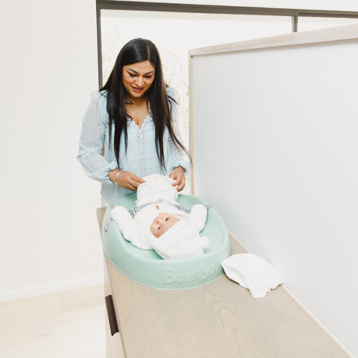 A woman in a light blue shirt is changing a baby wearing a white outfit and hat, who is lying in a mint green Baby Trend changing pad insert on a light wood table.