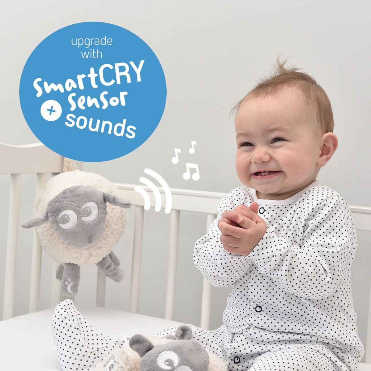 A baby in a white polka-dot sleeper sits in a crib next to a DELUXE Ewan the Dream Sheep, with a blue promotional badge reading 'upgrade with SmartCRY Sensor Sounds'.